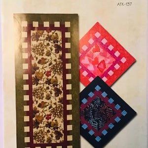 May include: A quilting pattern for a table runner and two placemats. The pattern is called "Waffle Time" and is from Atkinson Designs. The table runner features a green and brown floral design with a red and white checkered border. The placemats feature a red and pink heart design and a blue and black floral design.