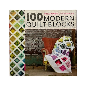 May include: A book titled "100 Modern Quilt Blocks" by Tula Pink. The book is next to a quilt draped over a red armchair. The quilt features a variety of colorful square patterns. The book's cover has a diamond pattern.