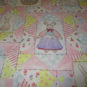 May include: A pink and white patchwork fabric with a repeating pattern of bunnies and floral designs. The fabric features a bunny wearing a purple dress and a basket of eggs.