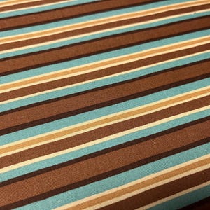 May include: Close-up of a fabric with a striped pattern. The stripes alternate between shades of brown, light blue, beige, and white. The fabric has a textured appearance, and the stripes run diagonally across the image.