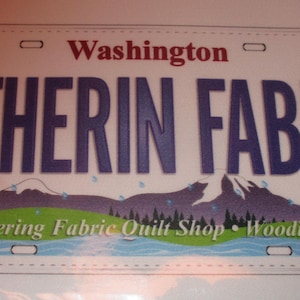 May include: A white and blue Washington state license plate with the text "GATHERIN FABRIC" and the text "Gathering Fabric Quilt Shop - Woodinville" below it.