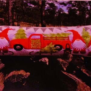 May include: A red truck with a white "Tree Farm" sign on the side is carrying a Christmas tree. Three gnomes are standing on the truck bed, each wearing a red hat and a white beard. The truck is on a pillow with a white background and a pattern of black triangles.