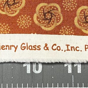 May include: Close-up of fabric with a repeating floral pattern in shades of brown and tan. A white strip across the fabric displays the text "Helping Hands" by Kim Diehl for Henry Glass & Co., Inc. Pattern 6875 www.henryglassfabrics.com. The fabric is placed on a ruler.