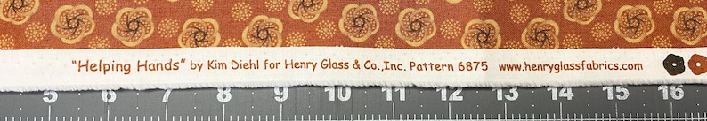 May include: Close-up of fabric with a repeating floral pattern in shades of brown and tan. A white strip across the fabric displays the text "Helping Hands" by Kim Diehl for Henry Glass & Co., Inc. Pattern 6875 www.henryglassfabrics.com. The fabric is placed on a ruler.