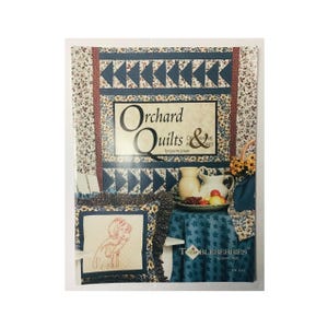 May include: A book titled "Orchard Quilts & Dreams & Accessories" by Lynette Jensen. The cover features a patchwork quilt with a blue and white floral pattern, a red and white floral pattern, and a blue and white geometric pattern. The book is published by "The Tableberries" and has a picture of a girl holding an apple on the front cover.