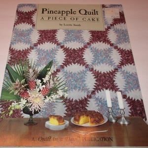 May include: A book titled "Pineapple Quilt: A Piece of Cake" by Loretta Smith. The cover features a quilt pattern with a pineapple motif and a floral arrangement in a vase. The book is a "Quilt in a Day" publication.