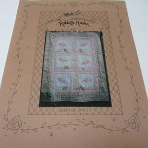 May include: A framed quilt with a light blue and pink color scheme. The quilt features a repeating pattern of squares with feather designs. The frame is tan with the words "Rabbit's Haven" and "Guardian Angels" written on it.