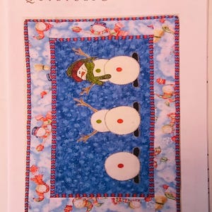 May include: A quilting pattern featuring three snowmen with red and white accents on a blue background with white snowflakes. The pattern is titled "Some Assembly Required" and has the number QC48.