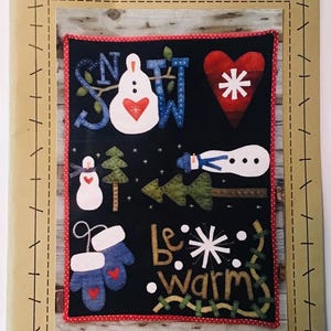 May include: A pattern for a quilted wall hanging with a winter theme. The design features a snowman, a heart, mittens, and the words "Snow" and "Be Warm". The pattern is titled "Snow Happy" and is from Wooden Spool Designs.