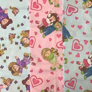 May include: Three fabric swatches with a repeating pattern of dolls and hearts. The fabric on the left is light blue with a white doll, the middle fabric is pink with a white doll, and the fabric on the right is light blue with a white doll.