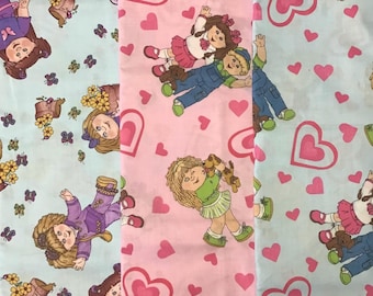 May include: Three fabric swatches with a repeating pattern of dolls and hearts. The fabric on the left is light blue with a white doll, the middle fabric is pink with a white doll, and the fabric on the right is light blue with a white doll.