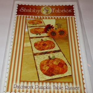 May include: A sewing pattern for a patchwork pumpkin table runner. The pattern features a design of four pumpkins in a row, with a sunflower design in the background. The pattern is titled "Shabby S*F Fabrics" and "Patchwork Pumpkin Table Runner".