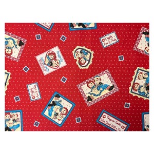 May include: Red fabric featuring Raggedy Ann and Andy illustrations in various shapes. The fabric includes rectangular and heart-shaped designs with the text "Ann & Andy" and "Things are Sweeter When Shared With a Friend."