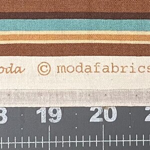 May include: Fabric sample with brown, teal, and beige horizontal stripes. The text "JAVA by DEB STRAIN for moda" is printed on a white strip, along with other details. The fabric is 100% cotton.