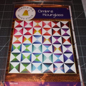 May include: A quilting pattern for an ombre hourglass quilt. The pattern is printed on a white background with a colorful quilt design. The pattern is titled "Ombre Hourglass" and is from the company "Maywood Studio".