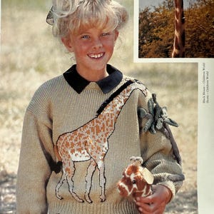 Puede incluir: Un suéter beige con un diseño de jirafa, usado por una persona. La jirafa es marrón y blanca, con un contorno negro. La persona también sostiene una jirafa de peluche. El suéter tiene un cuello negro.