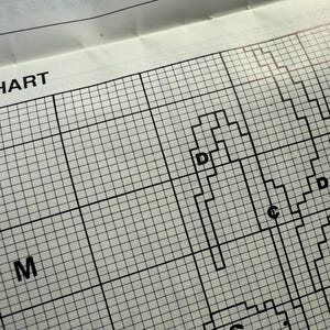 May include: Close-up of a cross-stitch pattern chart with a grid of squares. The chart features black lines forming shapes and letters, including "D", "C", and "M". The words "ER CHART" are visible at the top.