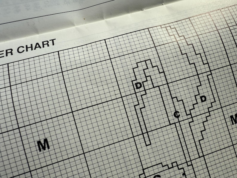 May include: Close-up of a cross-stitch pattern chart with a grid of squares. The chart features black lines forming shapes and letters, including "D", "C", and "M". The words "ER CHART" are visible at the top.