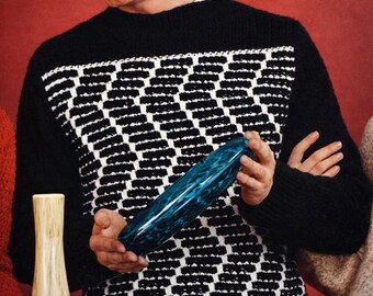 May include: A person holding a blue glass vase with a textured surface. The vase is long and oval-shaped. Other decorative vases are also visible. The person is wearing a black jumper with a white geometric pattern.