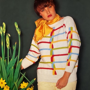May include: A white long-sleeved knit sweater featuring horizontal stripes in red, light blue, yellow, and green, with a textured weave. A bright yellow silk scarf is draped around the neck, and the sweater is paired with light yellow pants.