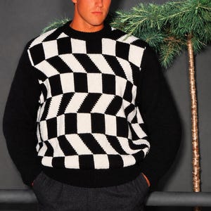 May include: A long-sleeved crew neck sweater featuring a black and white geometric checkerboard pattern on the front panel. The sleeves, collar, and hem are solid black. The knit design creates a three-dimensional optical illusion effect.