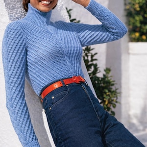 以下が含まれることがあります： テクスチャードニット模様のライトブルーのタートルネックセーター。セーターは濃い青色のジーンズと明るいオレンジ色のベルトと組み合わせています。赤いサングラスと赤いブレスレットを着用しています。