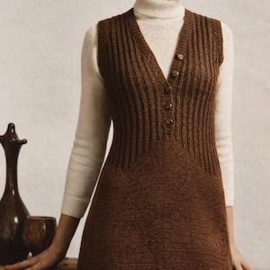May include: A brown knitted dress with a V-neck and button details. The dress is worn over a white turtleneck sweater. The dress has a ribbed texture on the bodice and a straight skirt. The overall style is vintage and classic.