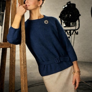 Könnte beinhalten: Ein marineblauer Pullover im Vintage-Stil mit Gürtel und dekorativer Brosche, kombiniert mit einem beigefarbenen Rock. Das Bild hat eine Retro-Ästhetik, mit einer Holzleiter und einem Studiolicht im Hintergrund. Der Stil erinnert an die 1960er Jahre.