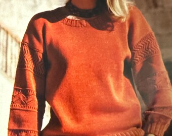 May include: A rust-coloured sweater with patterned sleeves. The sweater has a ribbed hem and cuffs. The sleeves feature a geometric design. The person is wearing a necklace and a skirt.