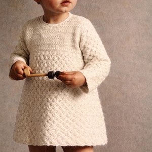 May include: A cream-colored knitted dress with long sleeves and a textured pattern. The dress is worn by a young child holding a wooden toy with black and brown accents. The background is a solid, neutral color.