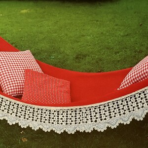 Puede incluir: Una hamaca roja con ribete de encaje blanco y tres almohadas. Dos almohadas tienen un patrón de cuadros rojos y blancos, y una tiene un patrón de lunares rojos. La hamaca está sobre un césped verde.