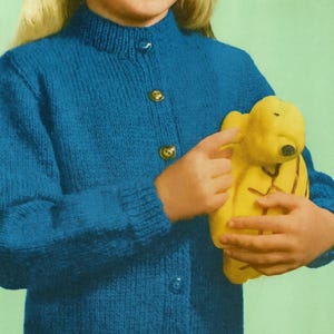 May include: A blue knitted cardigan with gold buttons. The cardigan is worn by a person holding a yellow stuffed animal. The stuffed animal has a black nose and is wearing a yellow outfit. The background is a solid green colour.