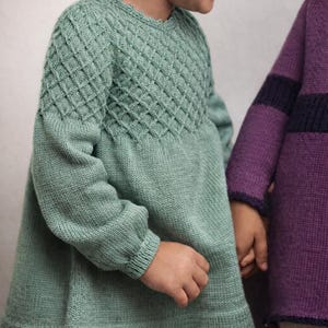 May include: A child's mint green knit dress with a diamond pattern on the bodice and long sleeves. The dress has a flared skirt and is made of soft, comfortable material. Another child wears a purple sweater.