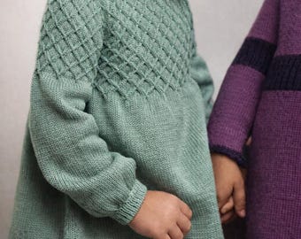 May include: A child's mint green knitted dress with a diamond pattern on the bodice and long sleeves. The dress has a flared skirt. Another child wears a purple sweater. The dresses are made of soft, comfortable material.