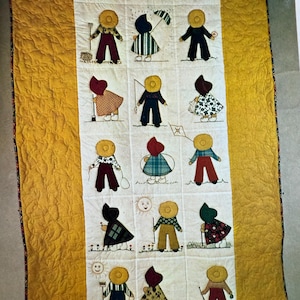 Puede incluir: Colcha hecha a mano con figuras aplicadas en varios atuendos, cada una con un sombrero amarillo. Las figuras est&aacute;n dispuestas en una cuadr&iacute;cula sobre un fondo blanco, bordeada por una tela amarilla texturizada. La colcha muestra un estilo de arte popular.