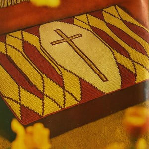 Może przedstawiać: Prostokątny gobelin z centralnym motywem krzyża. Krzyż jest brązowy na kremowym tle. Obramowanie przedstawia powtarzający się wzór żółtych i bordowych rombów. Idealny do dekoracji religijnych.