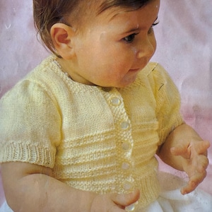 May include: A light yellow knitted baby cardigan with short sleeves and a button-up front. The cardigan has a ribbed texture on the sleeves and body, and is worn over a white garment. The background is a soft pink.