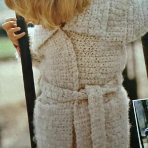 May include: A cream-colored, belted crochet coat with a large turn-down collar. The coat has a textured appearance and a matching belt. The coat is worn over red pants.