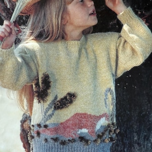 Könnte beinhalten: Ein hellgelber Strickpullover mit einem Landschaftsdesign mit einem roten Tier, grauen Pflanzen und einem blauen Sockel. Der Pullover hat lange Ärmel und einen runden Ausschnitt. Die Person auf dem Bild trägt einen Strohhut mit rotem Band.