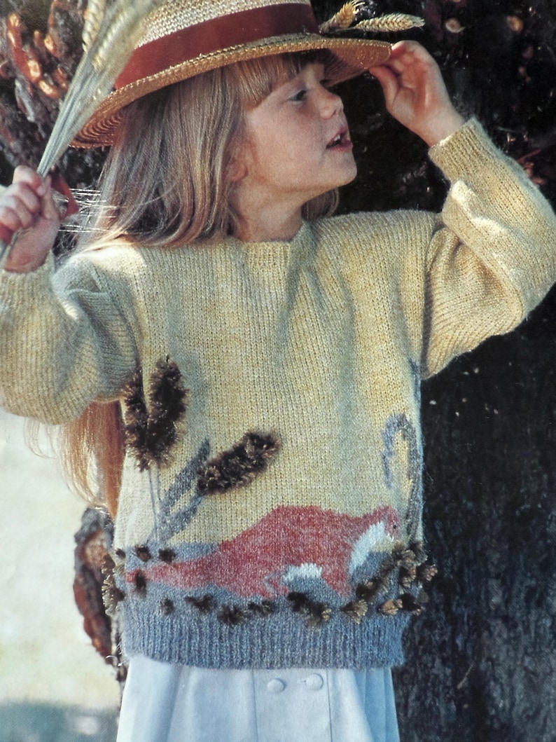 May include: A light yellow sweater with a landscape design featuring a red animal, gray and brown plants, and a gray border. The sweater has long sleeves and a round neckline. The person in the image is wearing a straw hat with a red band.