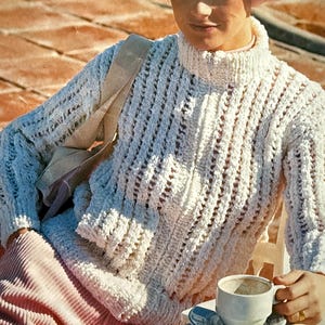 May include: A woman in a white textured high-neck sweater and a pink beret, seated outdoors. She holds a white teacup and saucer. The sweater features an open-knit design. The background includes a light-colored bag and pink corduroy trousers.
