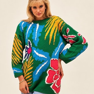 May include: A long-sleeve green knit sweater featuring a vibrant, multi-colored botanical pattern. The design includes large, stylized flowers and leaves in shades of bright pink, yellow, white, and blue, covering the front and sleeves.