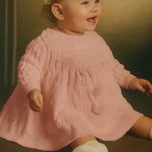 May include: A baby wearing a light pink knitted dress with a round neckline and long sleeves. The dress has a textured cable knit pattern on the sleeves and bodice, with a gathered skirt. The baby is also wearing white socks and a white shoe.