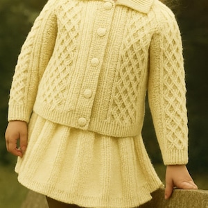 May include: A light yellow knitted cardigan and skirt set. The cardigan has a collared neckline and button closure. The skirt is pleated. The knitwear features a diamond pattern. The outfit is suitable for a child.