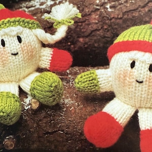 May include: Two handmade knitted elf dolls. Each doll has a white body, rosy cheeks, and a smiling face. They wear red and green hats and mittens. One elf has a white pom-pom. The dolls are set against a brown log background.