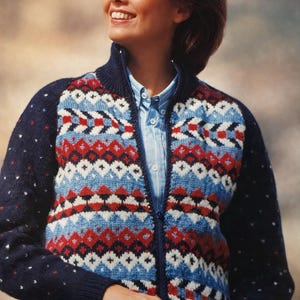 May include: A navy blue zip-up sweater with a geometric pattern in red, white, and light blue. The sweater has a high collar and is worn over a light blue button-down shirt. The sleeves are navy blue with small white dots.