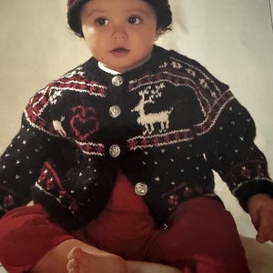 May include: A baby wearing a black knit hat with red tassels and a matching black cardigan. The cardigan features a white reindeer design and red accents. The baby is also wearing red pants.