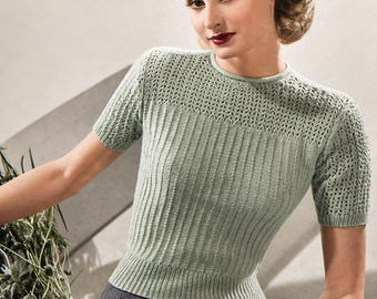 May include: A pale green short-sleeved knitted top with a ribbed design. The top features a crew neck and a patterned knit detail at the upper chest and sleeves. It is worn with a dark grey skirt.