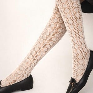 Puede incluir: Medias altas hasta el muslo de color crema con estampado de diamantes, combinadas con mocasines de cuero negro. Las medias tienen un diseño de tejido abierto y delicado. Los mocasines presentan una hebilla metálica dorada y una puntera.