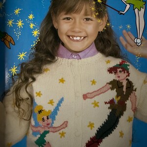 May include: A young person wearing a cream-colored cardigan sweater featuring embroidered images of Peter Pan and Tinkerbell. The sweater has yellow star accents and a button-up front. The background includes the words "Peter Pan" and illustrations.